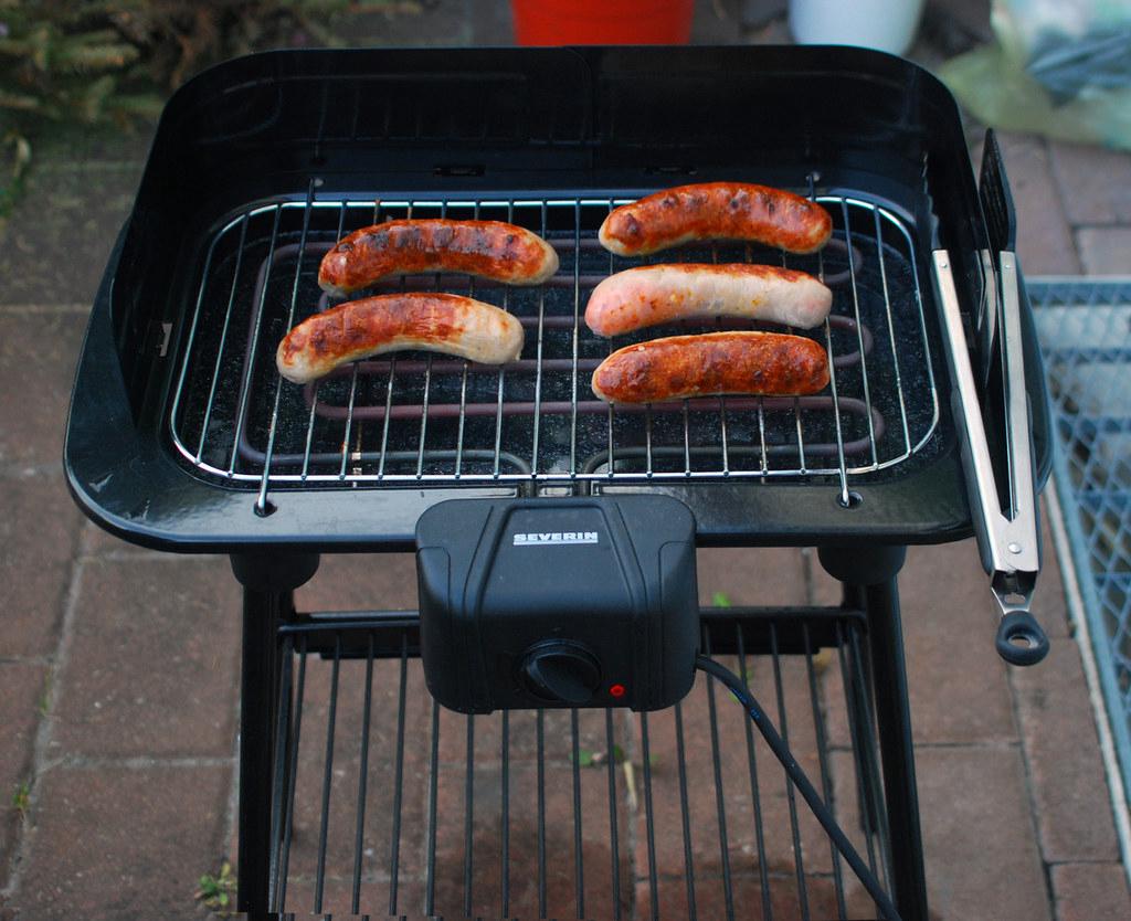 Ein Elektrogrill auf einem Holztisch im Garten, umgeben von grünen Pflanzen